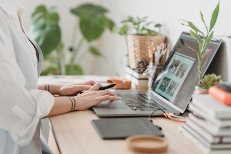 side view of a woman’s hands on a laptop computer keyboard. There are books, plants, computer peripherals, and a jar of pens and pencils on the desk.